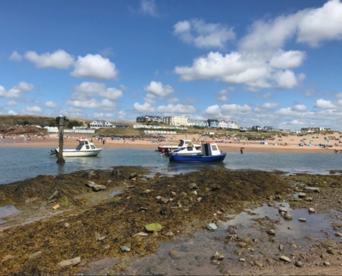 Image is looking across the sea to a sandy shore, golden sand, small boats in the foreground on a blue sky, sunny day