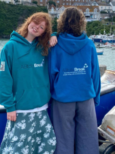 Two teenagers standing in a harbour, both wearing A Bit of a Break hoodies in cobalt blue and teal. One with long wavy red hair is smiling at the camera, leaning on the other teenager whose back is to the camera