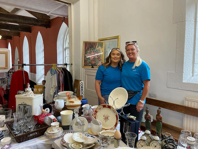Two women standing in a church hall wearing A bit of a Break blue t shirts, a table of crockery in front of them. They're smiling at the camera, one has long blonde hair in a pony tail and sunglasses on her head, the other with red wavy shoulder length hair. They're both smiling at the camera.