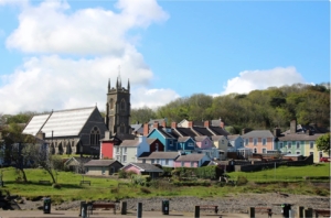 Image is of a traditinal dark grey stone church with a steeple and rows of coloured cottages, set amongst greenery and a blue sky with the odd white cloud.