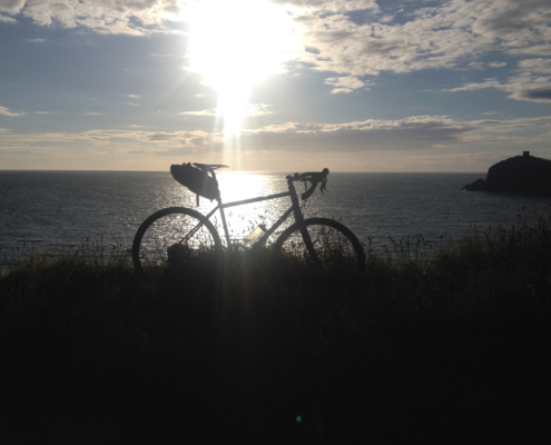 A bicycle on a grassy clifftop edge, silhouetted against an inky blue sea, white clouds and a lowering sun