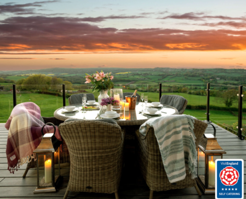 Six comfortable looking wicker armchairs covered in pretty check and striped blankets on some decking over looking the sun setting over green hills and rolling countryside. The table is covered with candles and cups and saucers, an ice bucket and champagne bottes and glasses