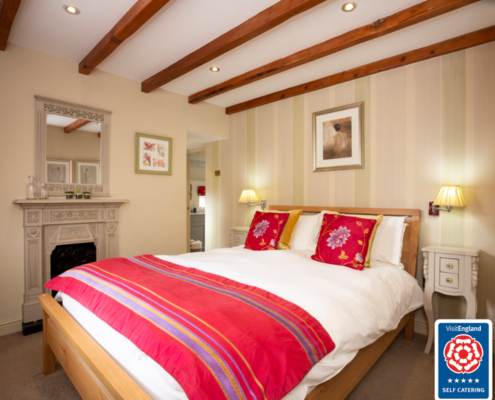 A cosy looking bedroom with a whitewashed ceiling and beams. White linen covered bed has pretty bright pink cushins and a thow. The room has a pretty pale grey fire surround with mirror above.